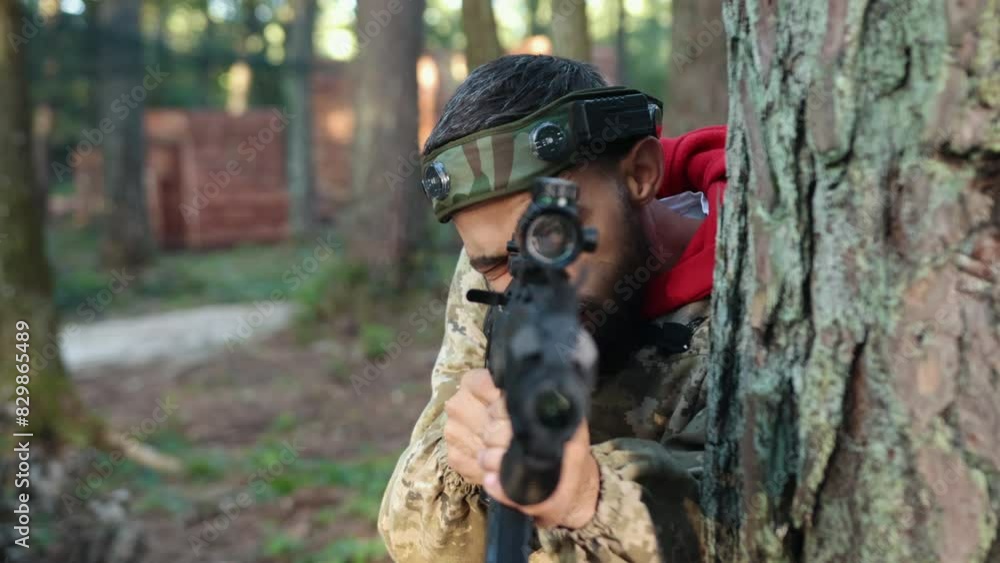 Camo gear, laser aim, tactical position. In forest setting, man outfitted in camo gear with marker on his head is propped against tree, cautiously aiming Laser tag gun and striking adversary. Camo gear, laser aim, tactical position. In forest setting, man outfitted in camo gear with marker on his head is propped against tree, cautiously aiming Laser tag gun and striking adversary.