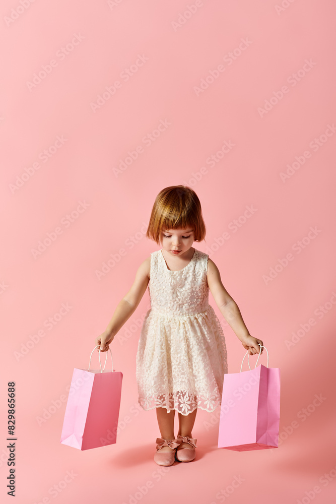 A little girl in a white dress happily holds shopping bags against a pink backdrop. A little girl in a white dress happily holds shopping bags against a pink backdrop.