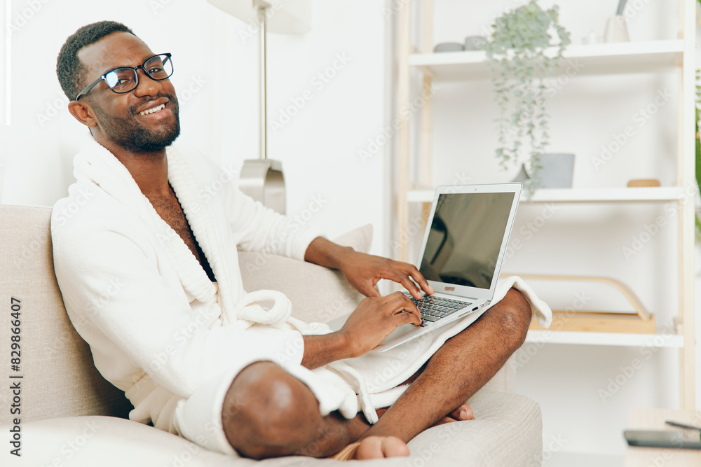 Smiling African American man in bathrobe working on a laptop on a modern sofa in the morning The cozy home environment, coupled with the use of technology and cyberspace, depicts a millennial Smiling African American man in bathrobe working on a laptop on a modern sofa in the morning The cozy home environment, coupled with the use of technology and cyberspace, depicts a millennial