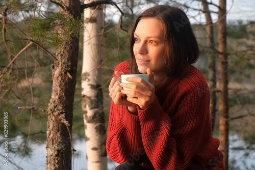Portrait relaxed woman drinking tea sitting on hill in forest mountain on nature. Tourist traveler, camper, hiker enjoying time outdoors. Female in red sweater enjoying woodland in camping in summer.