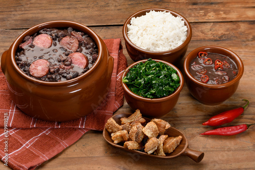 FEIJOADA IN A BROWN BOWL ON A WOODEN BASE, CABBAGE, RICE, BACON, PEPPER SAUCE