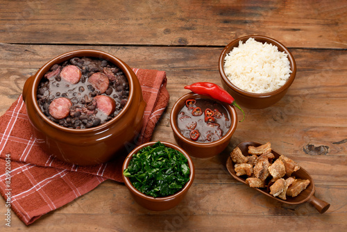 FEIJOADA IN A BROWN BOWL ON A WOODEN BASE, CABBAGE, RICE, BACON, PEPPER SAUCE
