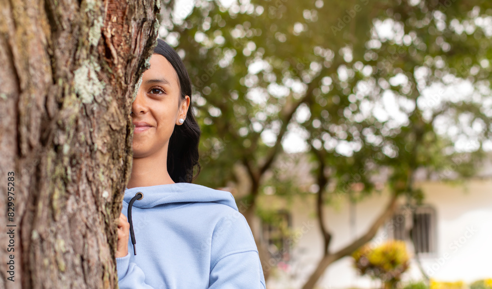 lifestyle: young happy latin woman smiling while playfully hiding behind a tree lifestyle: young happy latin woman smiling while playfully hiding behind a tree