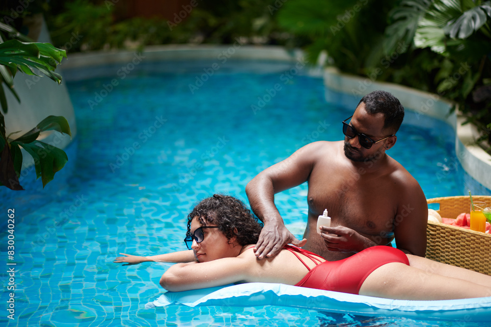 Young woman relaxing on air mattress on surface of blue water in swimming pool while her boyfriend applying lotion on her back