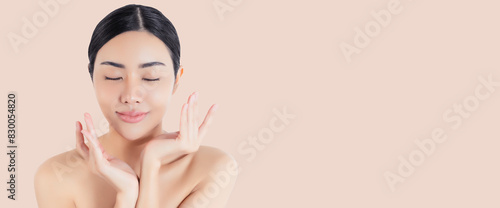 Young Asian woman with flawless skin gently touching her face, showcasing a natural beauty and skincare concept on a light beige background. Eyes closed with copy space.

