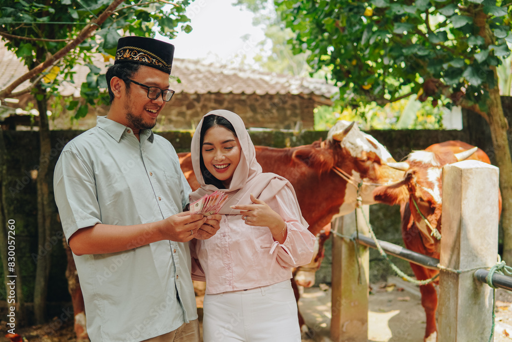 Excited young Asian Muslim couple pointing the money transaction they hold for cows cattle sacrifices. Eid Al Adha concept.