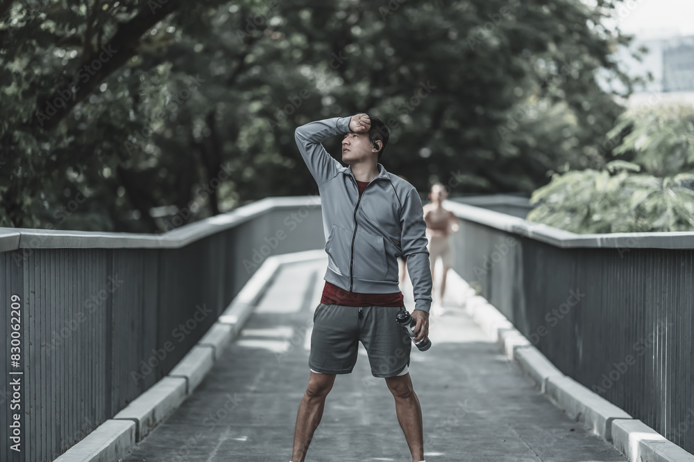 a tired young man wipe the sweat. Man resting, breathing and holding a bottle of drinking water on the road in the park morning. Health and Lifestyle in big city life concept. a tired young man wipe the sweat. Man resting, breathing and holding a bottle of drinking water on the road in the park morning. Health and Lifestyle in big city life concept.