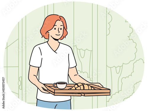 Woman wants to have breakfast in garden or in backyard of own house carries tray of hot tea and croissants. Happy girl carries delicious breakfast on tray to satisfy hunger before hard day at work.
