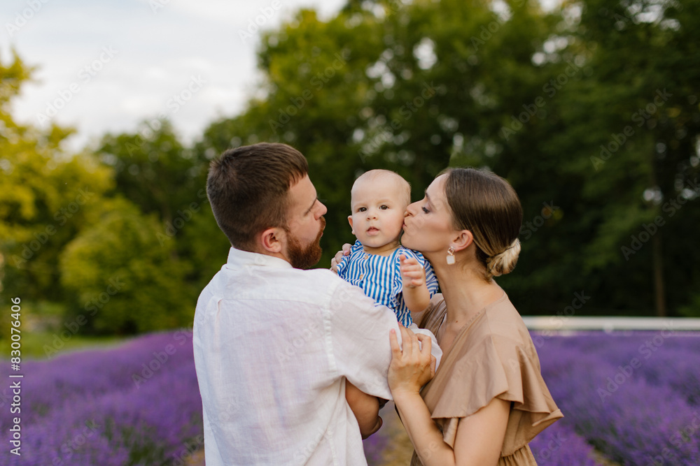 Fototapeta premium Family with a baby on a lavender field. A woman and a man with a beard are holding and kissing a newborn baby girl in a blue dress in a lavender field. Family, lifestyle concept.