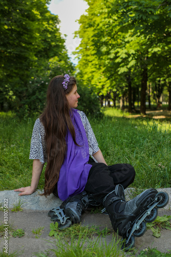 A young girl on roller skates and wearing a purple scarf sits on the grass in a city park in the spring.