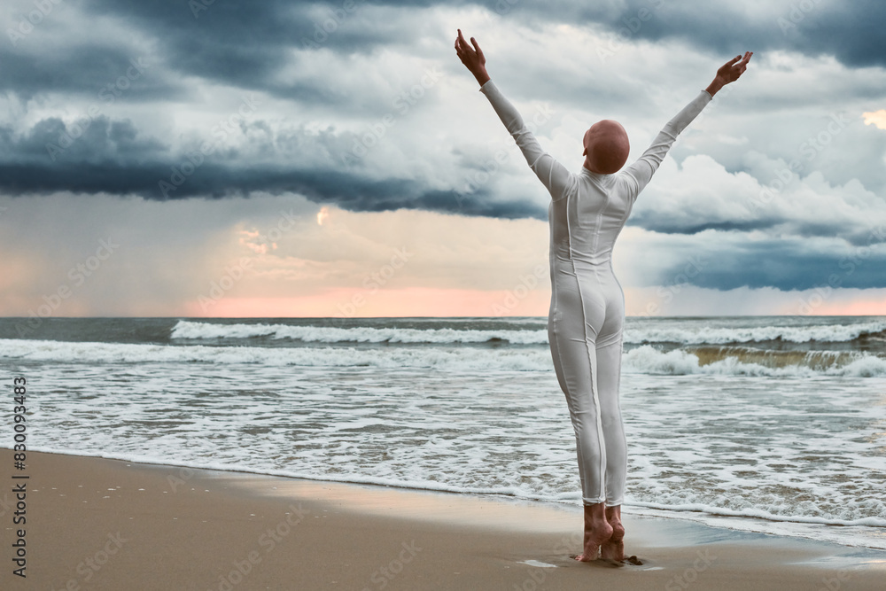 Hairless girl with alopecia in white futuristic suit standing on sea beach stretched out arms to cloudy sky, full length back view portrait, metaphoric performance with bald sensitive teenage girl Hairless girl with alopecia in white futuristic suit standing on sea beach stretched out arms to cloudy sky, full length back view portrait, metaphoric performance with bald sensitive teenage girl