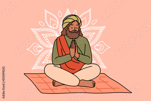 Meditating Indian man fakir prays sitting on mat with mandala sign behind back. Meditating adult guru with beard makes prayer gesture, performing esoteric and spiritualistic practices