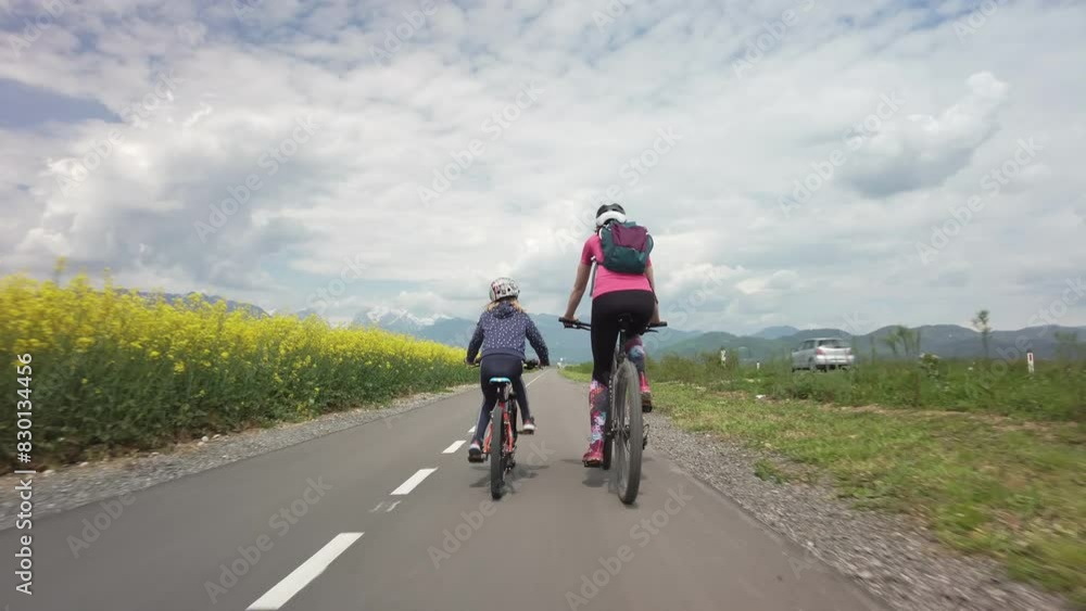 Mother and daughter, little girl, riding bikes along a scenic rural landscape on a summer cloudy day, rear view. Outdoors family activity concept. Mother and daughter, little girl, riding bikes along a scenic rural landscape on a summer cloudy day, rear view. Outdoors family activity concept.
