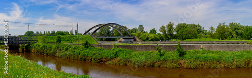 panorama photo of a beautiful landscape with lots of greenery, a river with an arched bridge for transport and a blue sky with clouds. © kristof Leffelaer