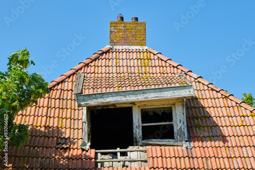 A dilapidated and sagging roof window on an orange roof of an abandoned farm in front of a blue sky. Concept of rural depopulation.