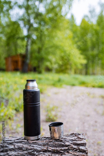 A glass bottle and a cup of drink are resting on a tree stump in the woods. Surrounded by grass and plants, they contrast with the natural wood and asphalt road surface nearby