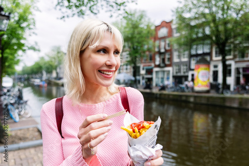 Photography Happy young tourist woman eating fries while enjoying summer vacation in Amsterdam, Netherlands