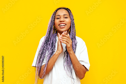 cheerful african american woman with colored dreadlocks applauding and congratulating on yellow isolated background, hipster girl with purple pigtails and unique hairstyle clapping and laughing