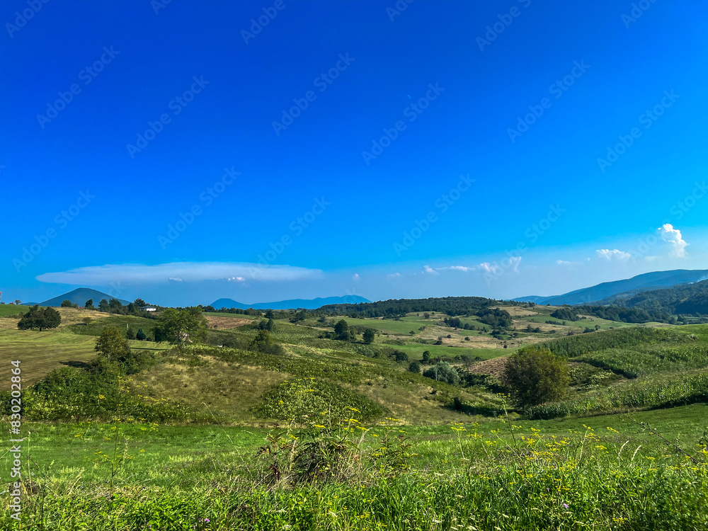 View of small hills and larger mountains with a small field and a house, Han Kola, Bosnia