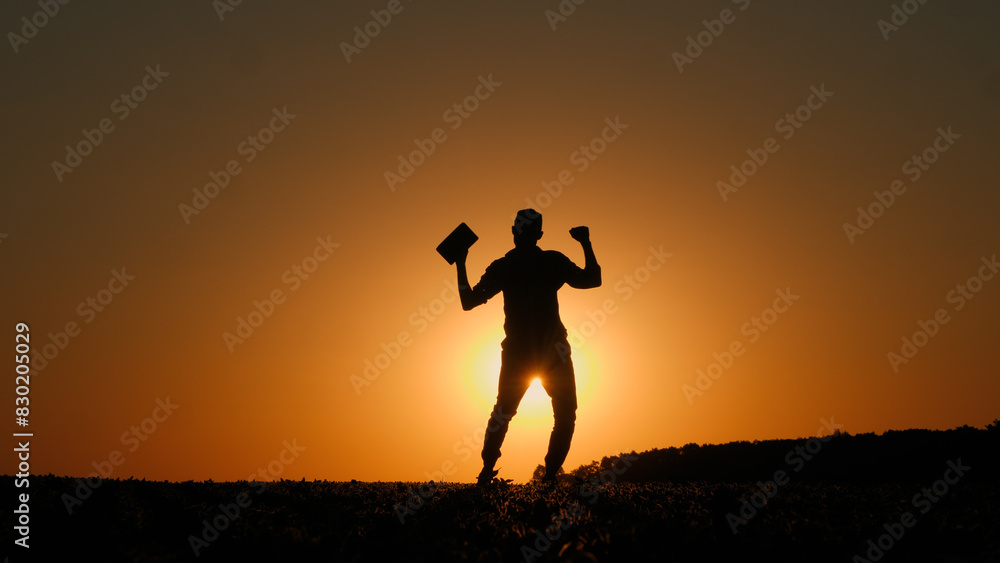 Fototapeta premium A man with a tablet in his hand dances a cheerful dance in a field at sunset