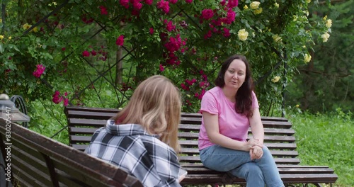 Mom talking with teen daughter sitting in summer park on benches among flowering trees. Mother looking at girl teenager explaining something. Family conversation outdoors, leisure, pastime together.