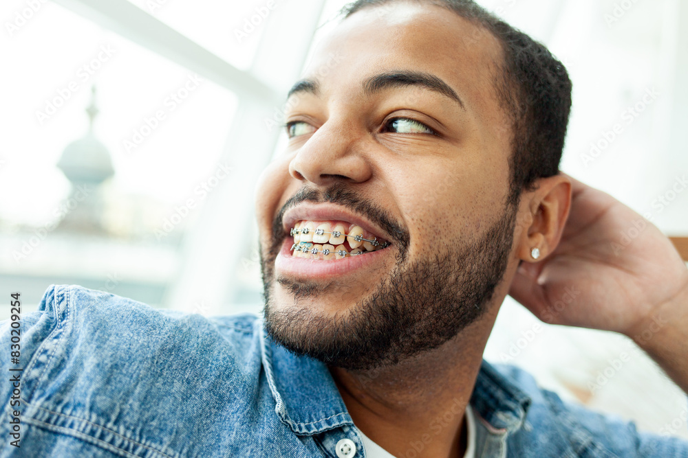 Obraz premium happy african american man with braces laughs and looks out the window in a white cafe, relaxed man in a denim shirt dreams and smiles