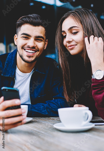 Cheerful teen hipster guy looking at camera while showing her girlfriend video on smartphone  romantic couple in love spending time together on cafe terrace viewing funny photos on mobile phone