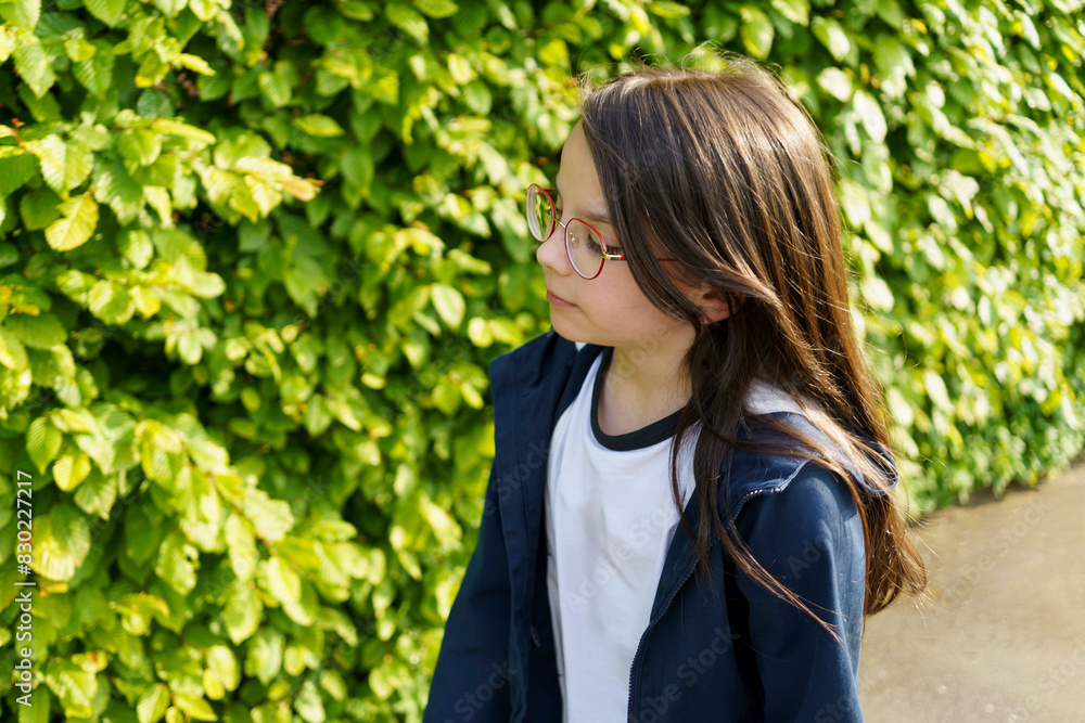 A little beautiful girl in red glasses stands against the background of a green hedge. A little beautiful girl in red glasses stands against the background of a green hedge.