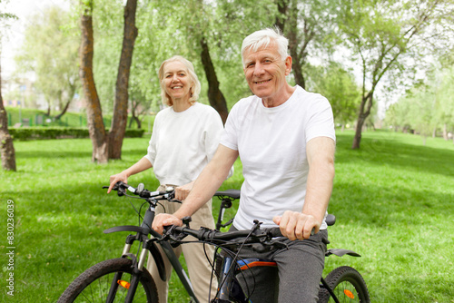 elderly senior couple rides bicycle in the park in the summer and smiles, old gray-haired man and woman are actively resting outdoors, old people practice cycling in forest
