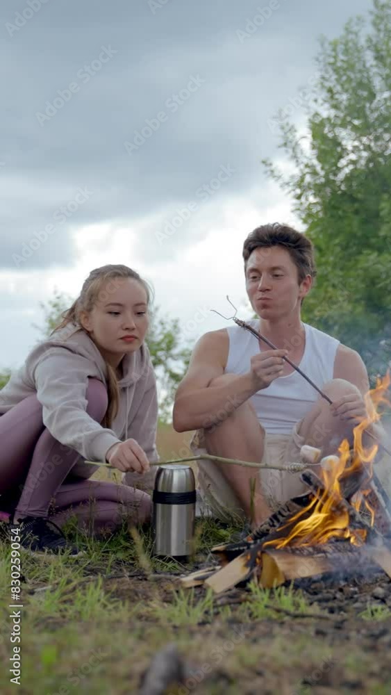 Young couple sits on mat near campfire frying marshmallows. Handsome man smiles and laughs looking at woman burnt down marshmallow on wooden stick Vertical Shot. Young couple sits on mat near campfire frying marshmallows. Handsome man smiles and laughs looking at woman burnt down marshmallow on wooden stick Vertical Shot.