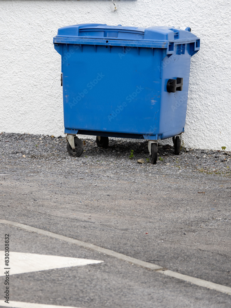 A blue trash can is sitting on the side of a road. The trash can is empty and has a white stripe on the bottom. White wall background. Rubbish removal in a city. A blue trash can is sitting on the side of a road. The trash can is empty and has a white stripe on the bottom. White wall background. Rubbish removal in a city.