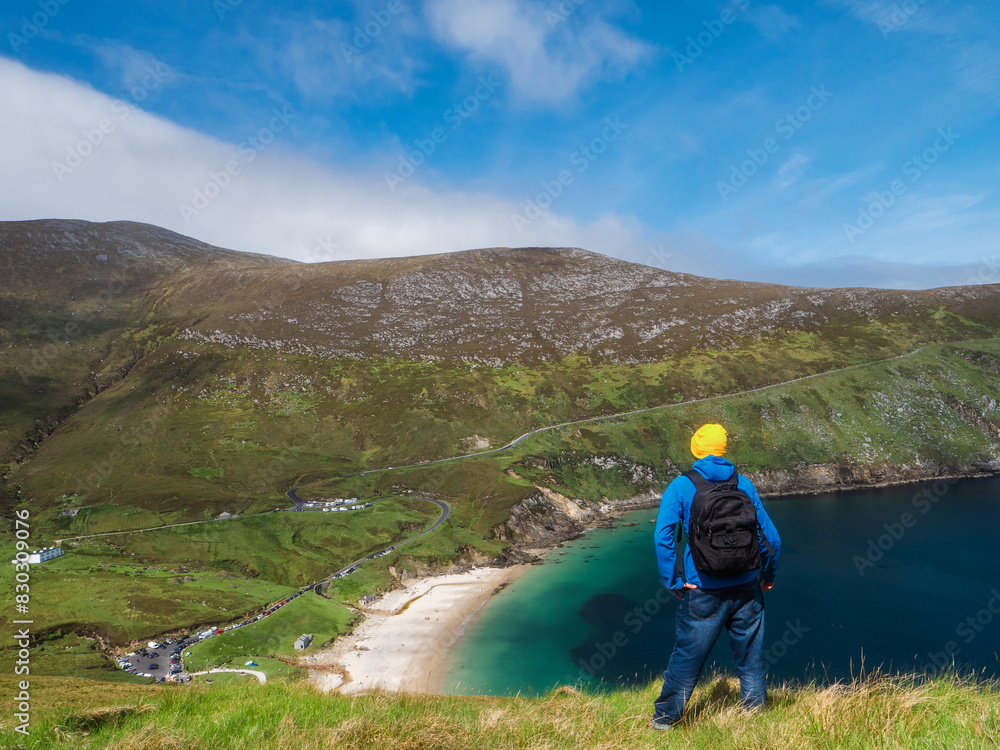A man in a blue jacket stands on a hill overlooking a beach and a ocean. The sky is clear and the sun is shining, creating a peaceful and serene atmosphere. Keem bay and beach Ireland. Travel concept