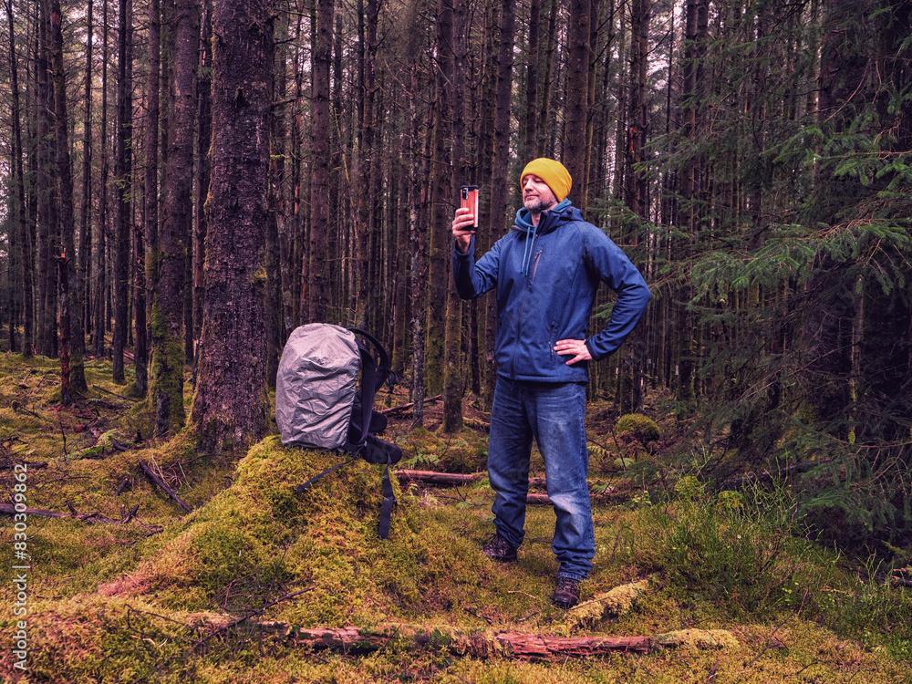 Naklejka premium Male tourist taking pictures on phone, his backpack on a tree stump. Scene is peaceful and serene, as the man is surrounded by nature. Travel and tourism theme. Model in blue jacket and yellow hat.
