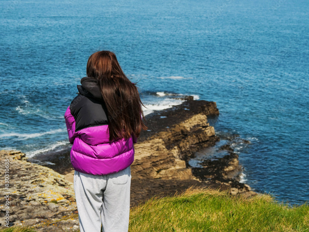 A girl in a purple jacket stands on stone shore. She is wearing gray sweatpants and. The ocean is in the background and the sky is cloudy and blue. Travel and tourism. Mullaghmore, Ireland.