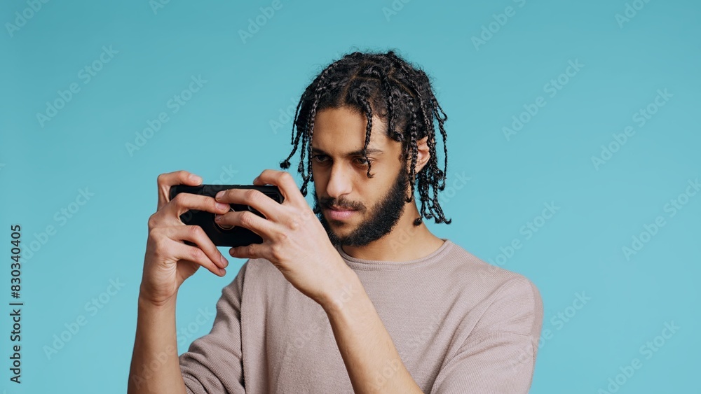 Gamer disappointed after receiving game over screen on cellphone touchscreen display. Middle Eastern man nodding head, upset after losing videogame, playing on phone, studio background, camera B Gamer disappointed after receiving game over screen on cellphone touchscreen display. Middle Eastern man nodding head, upset after losing videogame, playing on phone, studio background, camera B