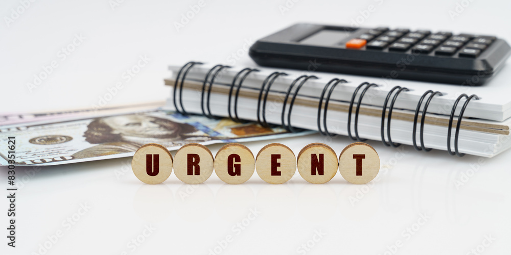 On a high surface lie a notepad, a calculator, dollars and wooden circles with the inscription - URGENT On a high surface lie a notepad, a calculator, dollars and wooden circles with the inscription - URGENT