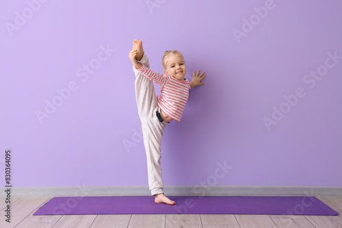 Cute little girl practicing yoga on mat against lilac background