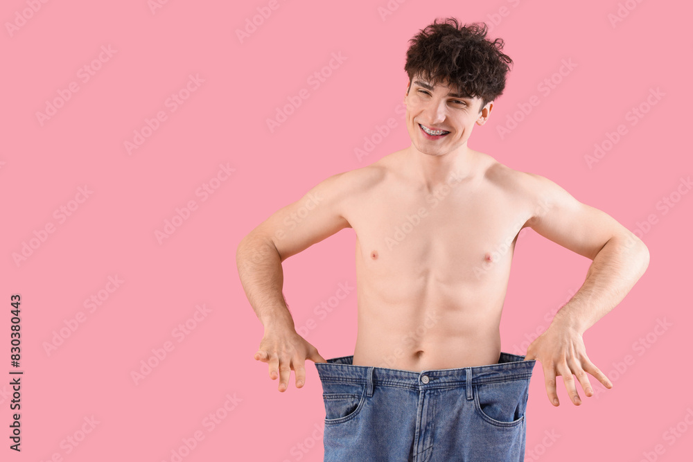 Handsome happy young sporty man in loose jeans on pink background. Weight loss concept Handsome happy young sporty man in loose jeans on pink background. Weight loss concept