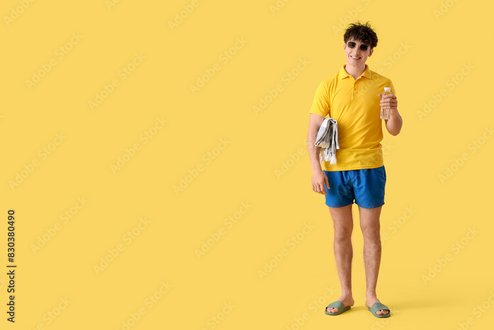 Fototapeta premium Handsome young happy man with beach blanket and bottle of water on yellow background
