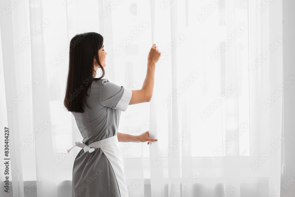 Young chambermaid adjusting window curtain in room Young chambermaid adjusting window curtain in room