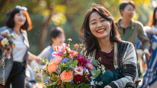 A smiling Asian woman in a wheelchair, holding a bouquet of flowers, surrounded by a diverse group of friends celebrating in a park