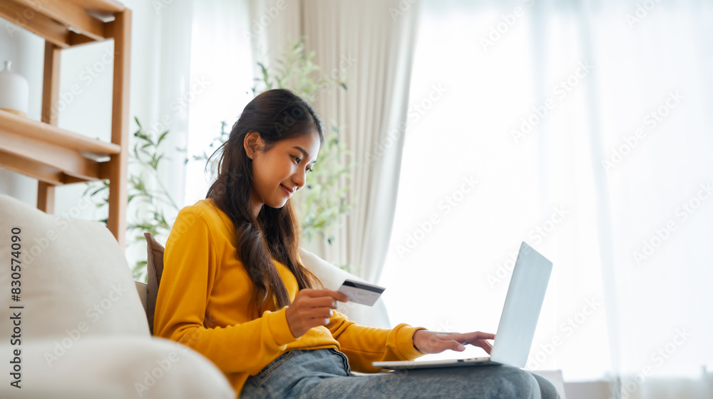 Young asian woman sitting on sofa in living room, makes online banking payments through the internet from bank card on computer laptop. Shopping online on notebook with credit card Young asian woman sitting on sofa in living room, makes online banking payments through the internet from bank card on computer laptop. Shopping online on notebook with credit card