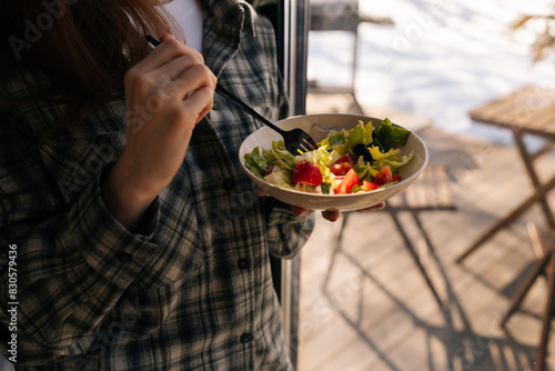 Cropped shot of unrecognizable healthy young woman holding salad bowl and eat clean vegetables for lunch in weight loss diet and wellness standing by window on sunny day. Healthy food concept.