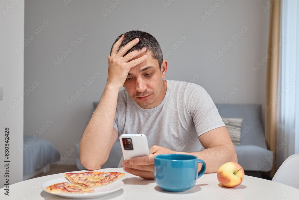 Unhappy sad Caucasian man wearing T-shirt sitting at table in home interior having troubles being overworked using mobile phone browsing web pages scrolling online Unhappy sad Caucasian man wearing T-shirt sitting at table in home interior having troubles being overworked using mobile phone browsing web pages scrolling online