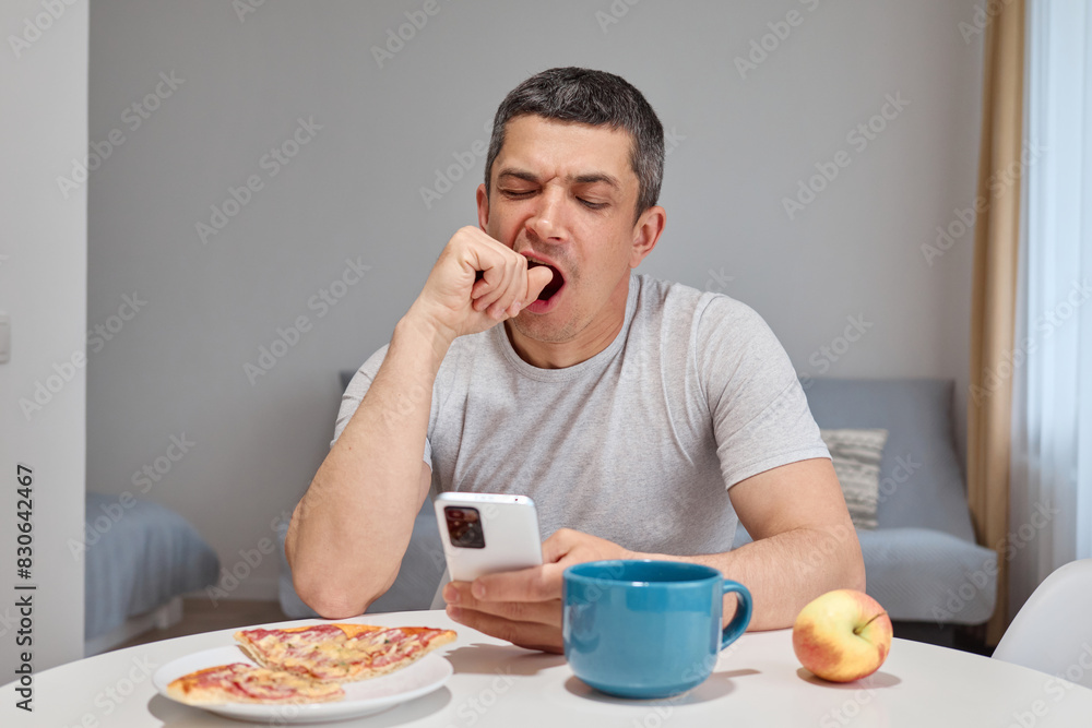 Tired sleepy Caucasian man wearing T-shirt using smartphone while having dinner yawning texting in social networks sitting at table in home interior Tired sleepy Caucasian man wearing T-shirt using smartphone while having dinner yawning texting in social networks sitting at table in home interior