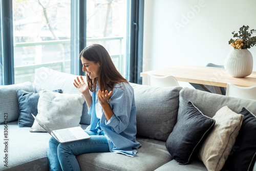 Young woman waves goodbye. Smiling female uses laptop, sitting on sofa at home, beautiful girl chatting online on social networks and laughing with friends .Technology and friendship concept.