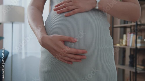 An expectant mother in a snug dress stands in contemplation, hands lovingly framing pregnant belly, highlighted by soft natural light.