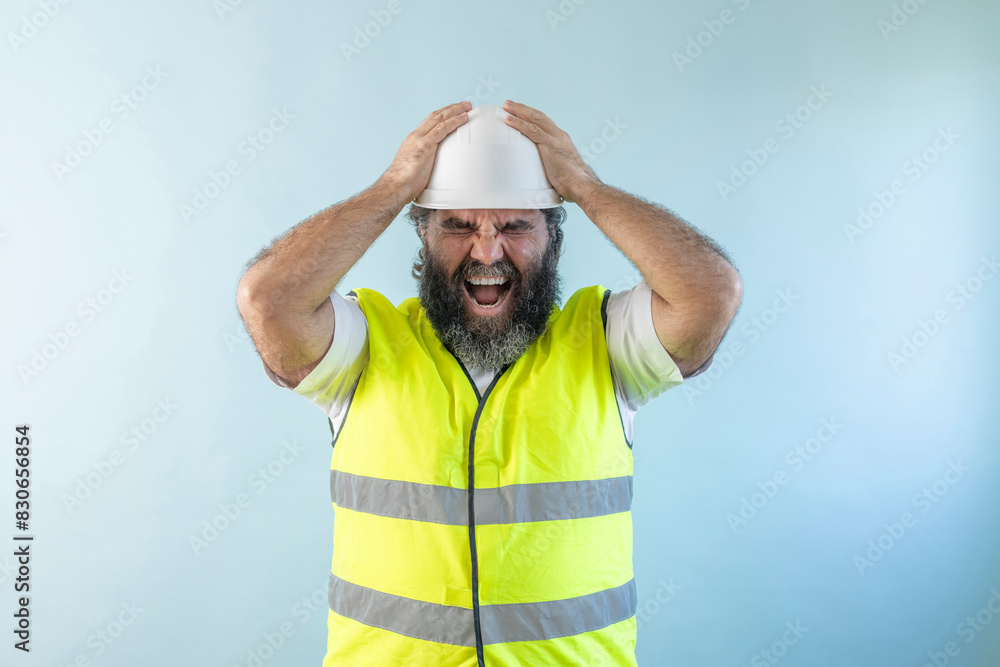 Naklejka premium stressed adult man engineer with beard and wearing helmet and safety vest, looking at camera on blue background holding his head