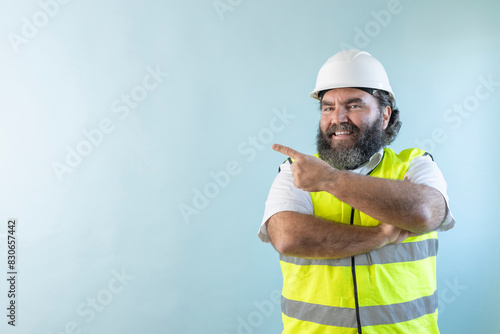 smiling adult man engineer with beard and wearing helmet and safety vest, looking at camera on blue background, with crossing arms
