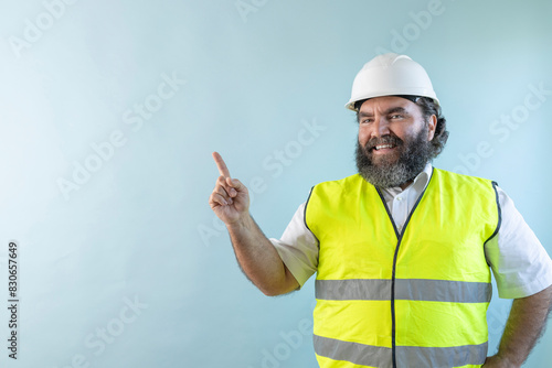 smiling adult man engineer with beard and wearing helmet and safety vest, looking at camera on blue background and pointing finger to the left